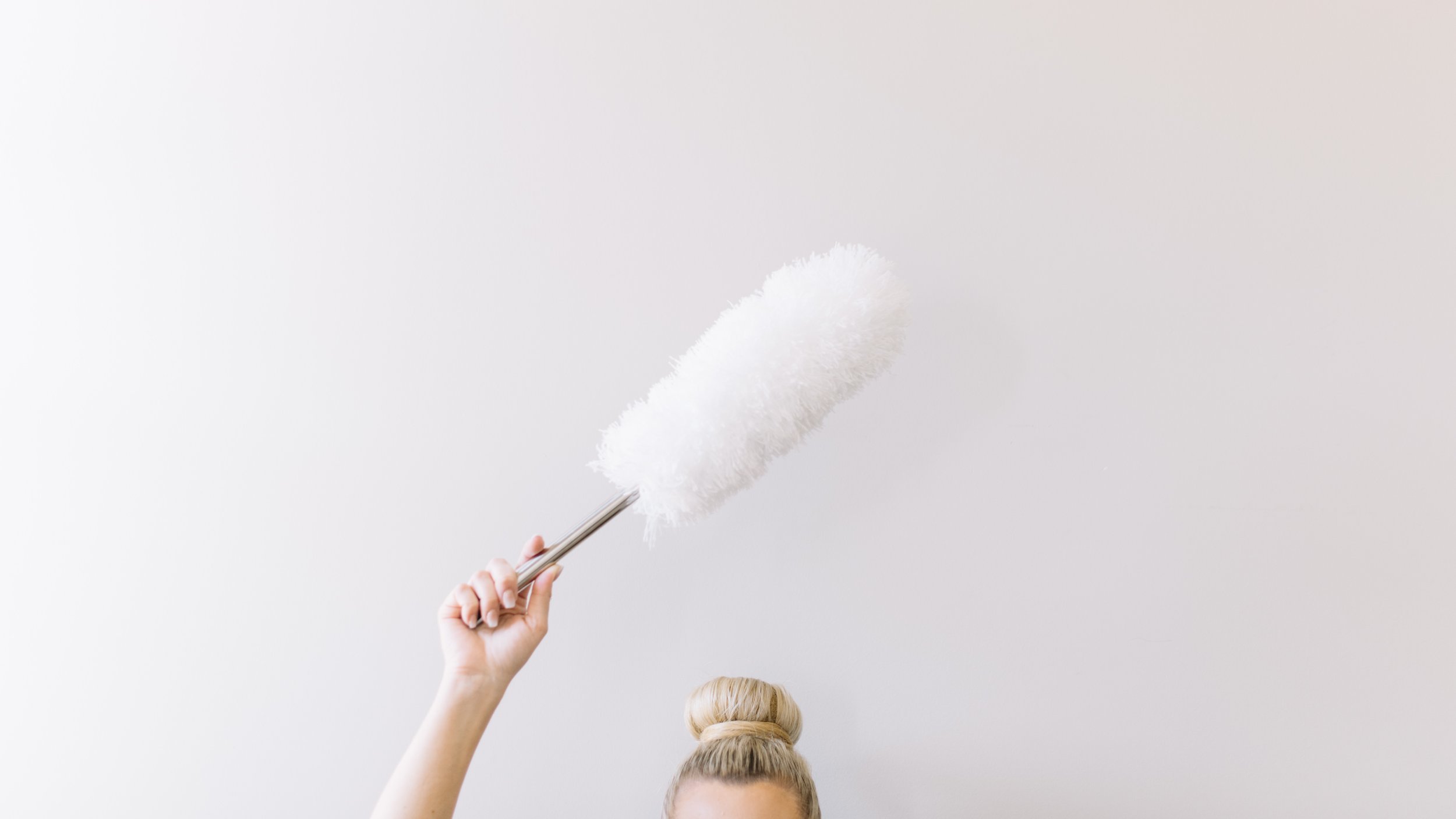A person's hand holding a fluffy white duster against a plain white wall